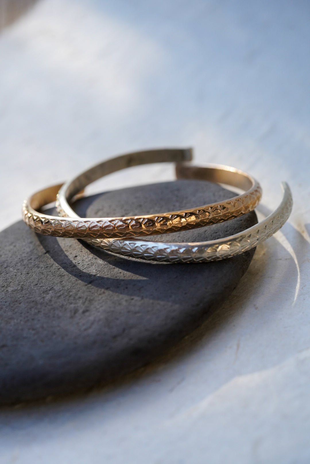 Two textured open cuff bracelets, one gold and one silver, displayed on a dark stone against a light gray background.