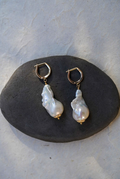 A pair of gold huggie earrings with large, irregular white baroque pearl drops, displayed on a dark gray stone against a light gray textured background.