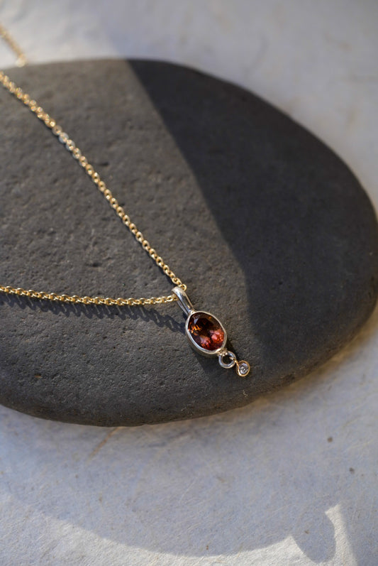 A delicate gold chain necklace with an oval amber-brown gemstone pendant and a small diamond accent stone, displayed on a dark gray stone slab against a light beige background.