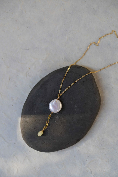 A gold chain necklace with a round iridescent white pearl pendant and a small gold teardrop opal, displayed on a dark stone over a light textured background.