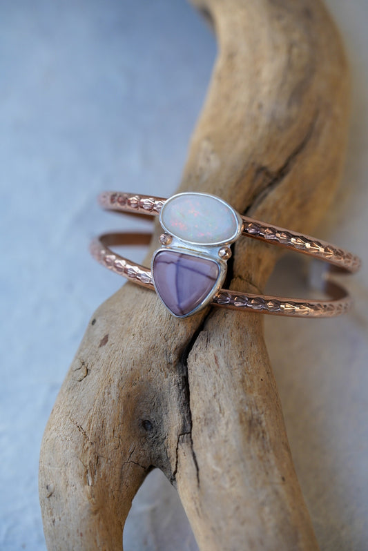 a detailed rose gold double band cuff displayed on a piece of driftwood. Set on the bands are an opal and jasper stone.