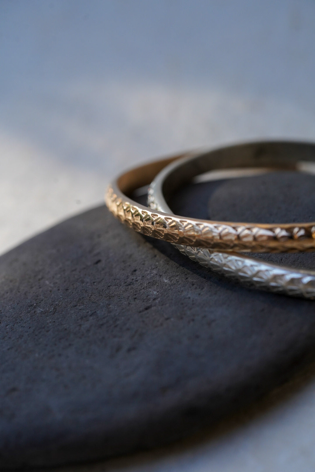 Two textured open cuff bracelets, one gold and one silver, displayed on a dark stone against a light gray background.