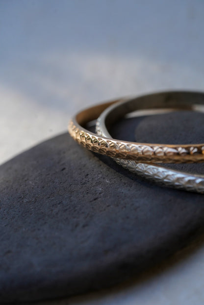 Two textured open cuff bracelets, one gold and one silver, displayed on a dark stone against a light gray background.