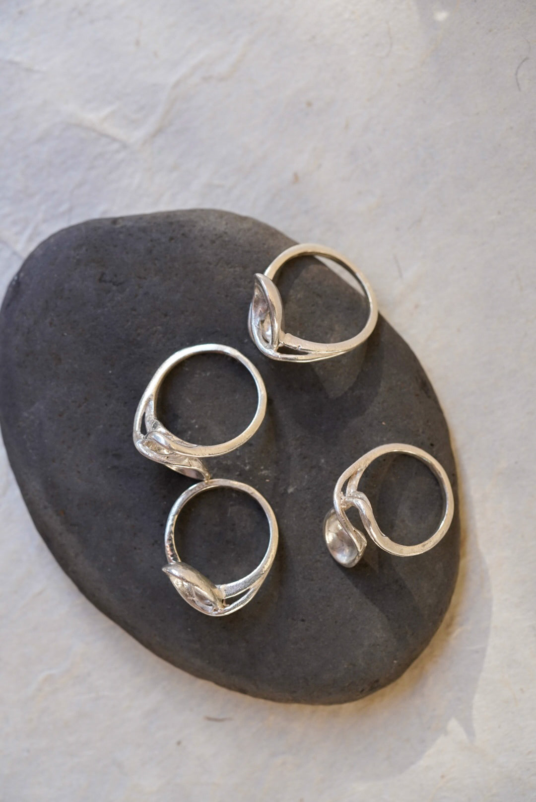Four silver rings with organic, flowing  wave-like band designs, arranged on a dark gray stone against a light textured background.