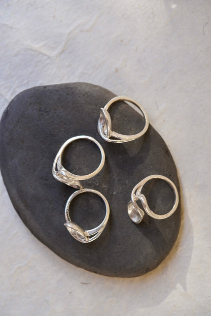 Four silver rings with organic, flowing  wave-like band designs, arranged on a dark gray stone against a light textured background.