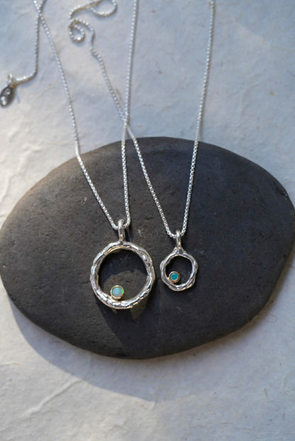 Two silver chain necklaces with open circular pendants, each featuring a small iridescent opal stones, displayed on a dark stone slab against a light textured background.