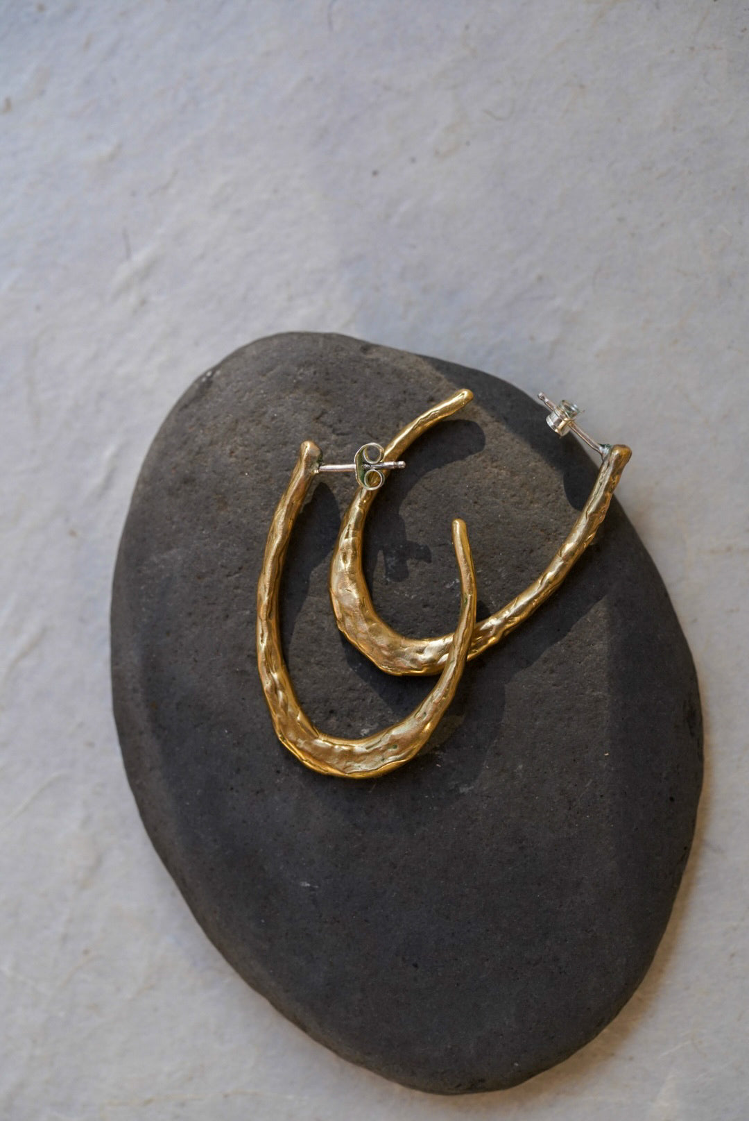 A pair of textured gold hoop earrings displayed on a dark gray stone against a light textured background.