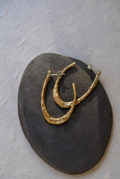A pair of textured gold hoop earrings displayed on a dark gray stone against a light textured background.