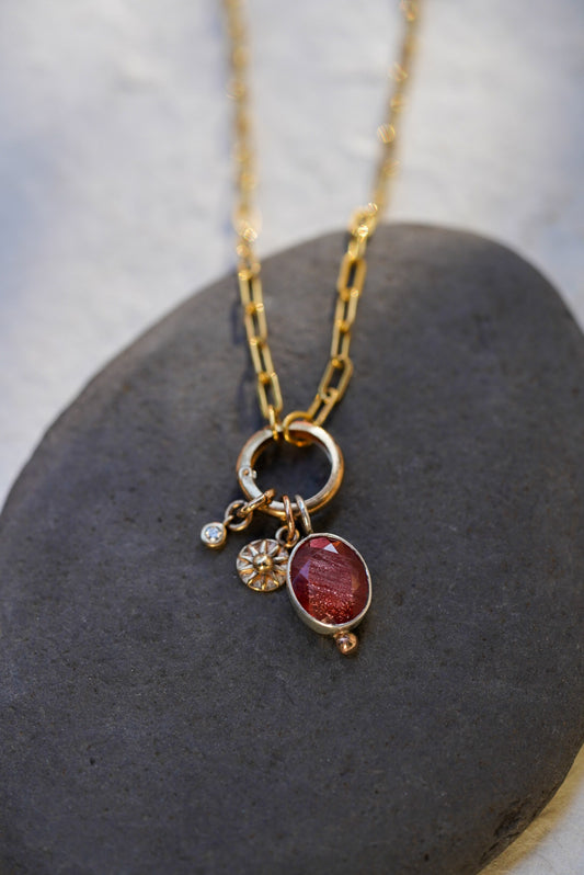 A gold chain necklace with a ring clasp and three dangling charms, including a faceted oval red sunstone, a sun and small Damond displayed on a dark gray stone against a light gray background.