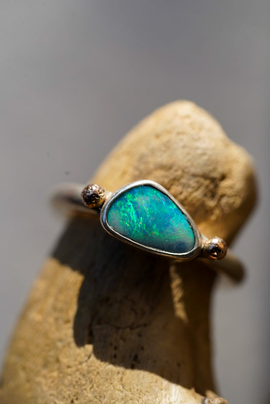 A close-up of a ring with a green-blue opal center stone and rose gold side accents, resting on a dark gray stone against a textured white background.