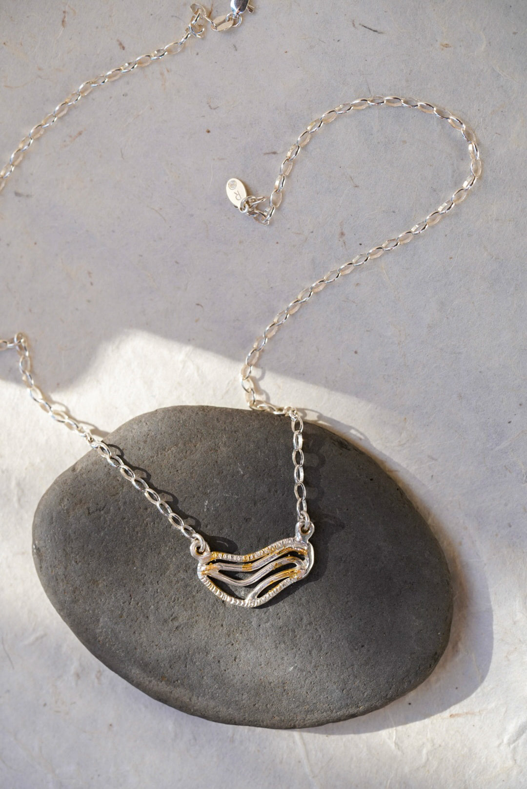 A silver chain necklace with organic shaped pendant featuring textured gold dusting, displayed on a smooth dark gray stone against a light gray background.