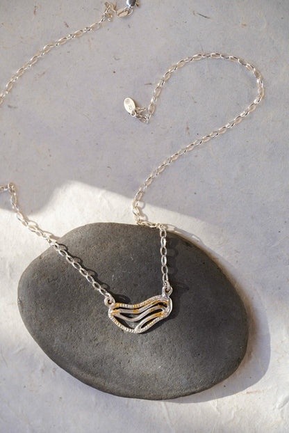 A silver chain necklace with organic shaped pendant featuring textured gold dusting, displayed on a smooth dark gray stone against a light gray background.