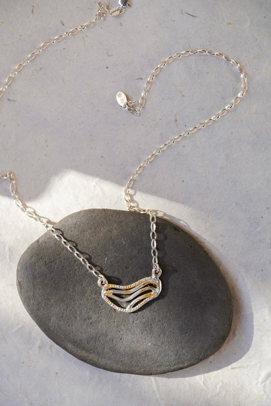 A silver chain necklace with organic shaped pendant featuring textured gold dusting, displayed on a smooth dark gray stone against a light gray background.
