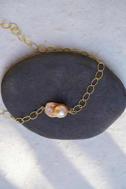 A gold chain necklace with a single irregular baroque pearl pendant, displayed on a dark gray stone against a textured off-white background.