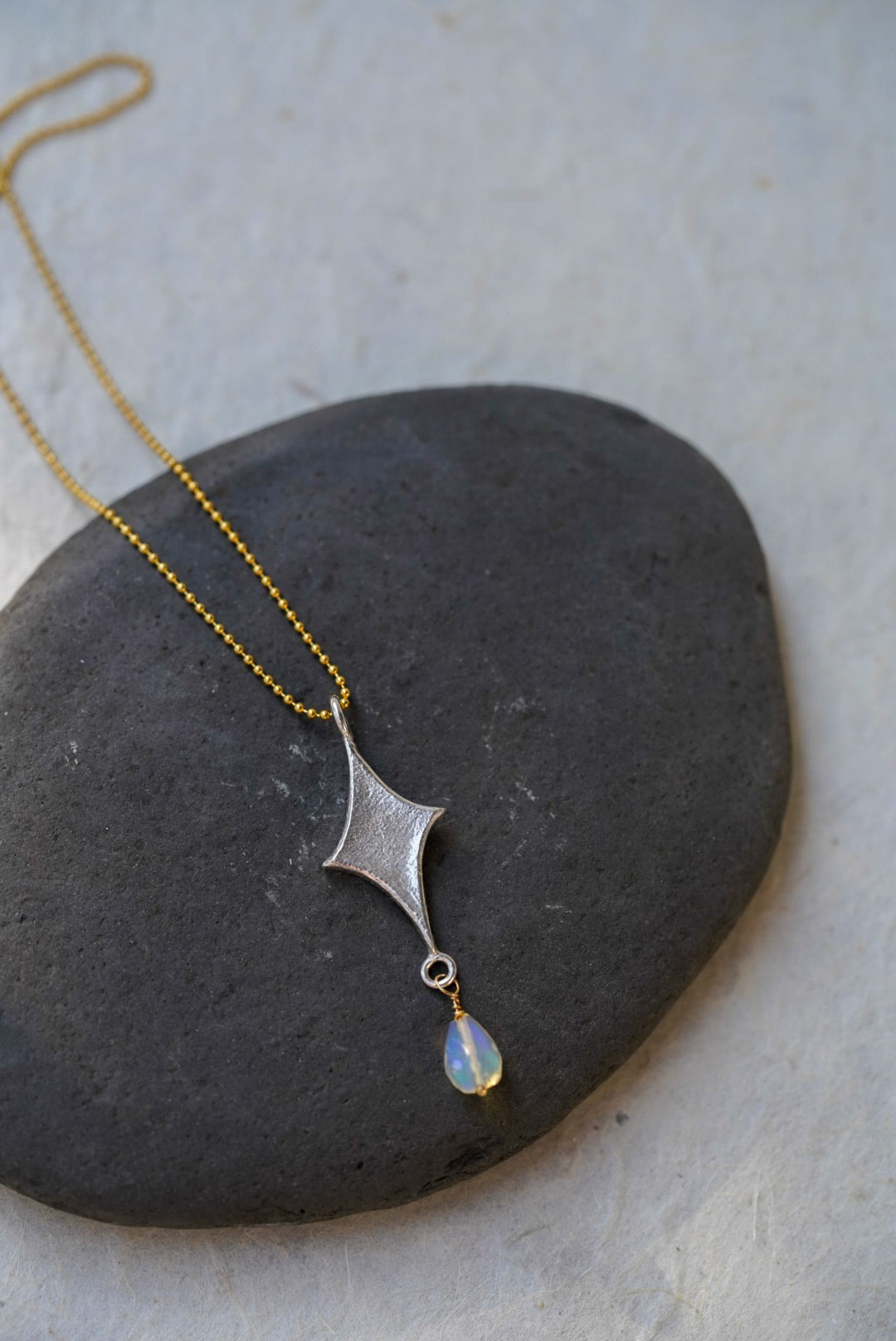 A delicate necklace with a textured silver starburst pendant and a small iridescent opal teardrop, resting on a dark gray stone against a light gray background.