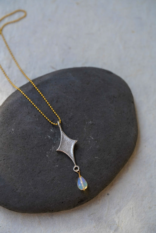 A delicate necklace with a textured silver starburst pendant and a small iridescent opal teardrop, resting on a dark gray stone against a light gray background.