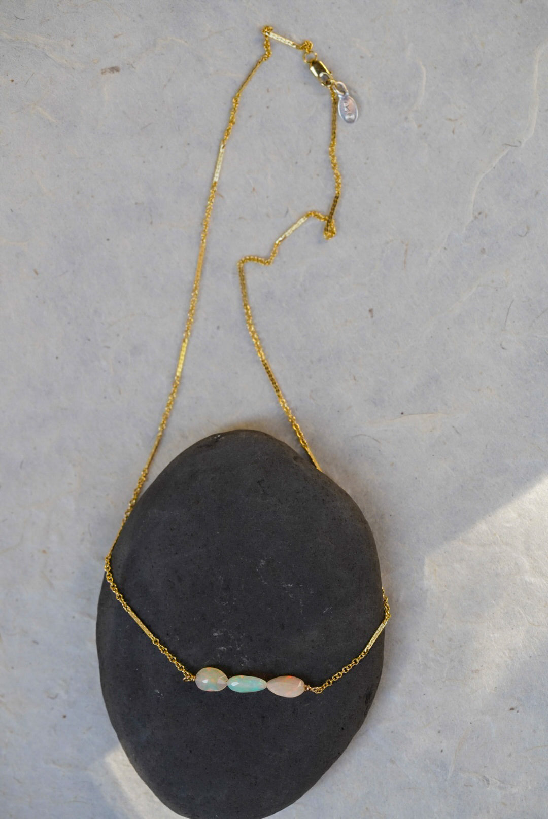 A delicate gold chain necklace draped over a dark gray stone, featuring three small opal beads in pale blue, pink, and white.