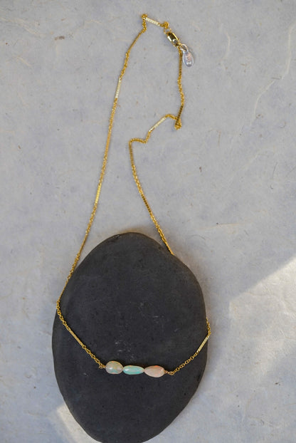 A delicate gold chain necklace draped over a dark gray stone, featuring three small opal beads in pale blue, pink, and white.