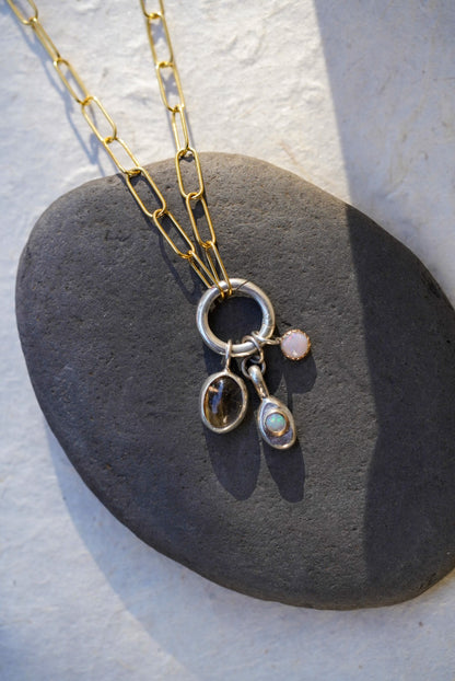 A gold chain necklace with a silver ring and three dangling charms, including a rutilated quartz and small opal and a small round pink opal stone, displayed on a dark gray stone against a light background.
