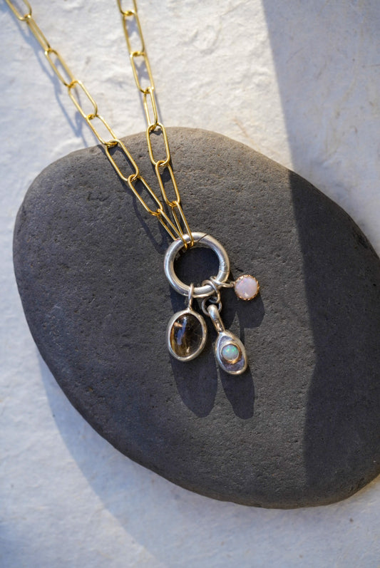 A gold chain necklace with a silver ring and three dangling charms, including a rutilated quartz and small opal and a small round pink opal stone, displayed on a dark gray stone against a light background.