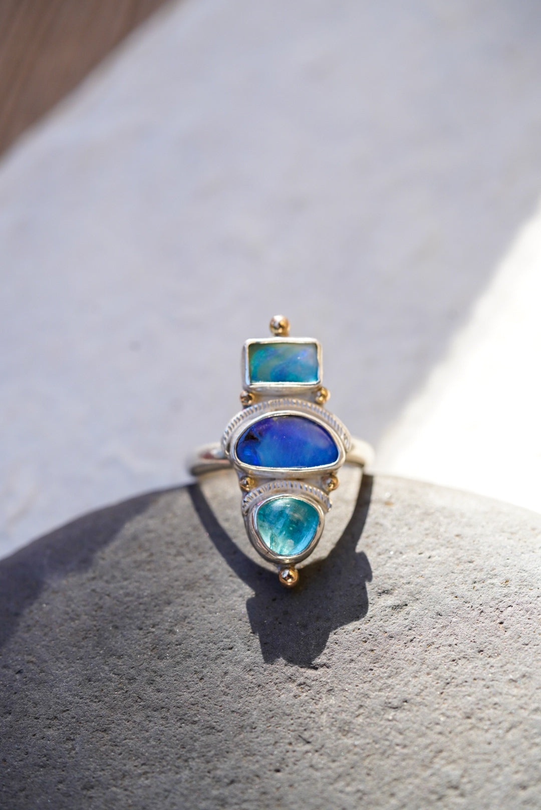A silver ring with three stacked bright blue stones in vibrant blue and turquoise tones, displayed on a piece of rock  against a neutral light background.