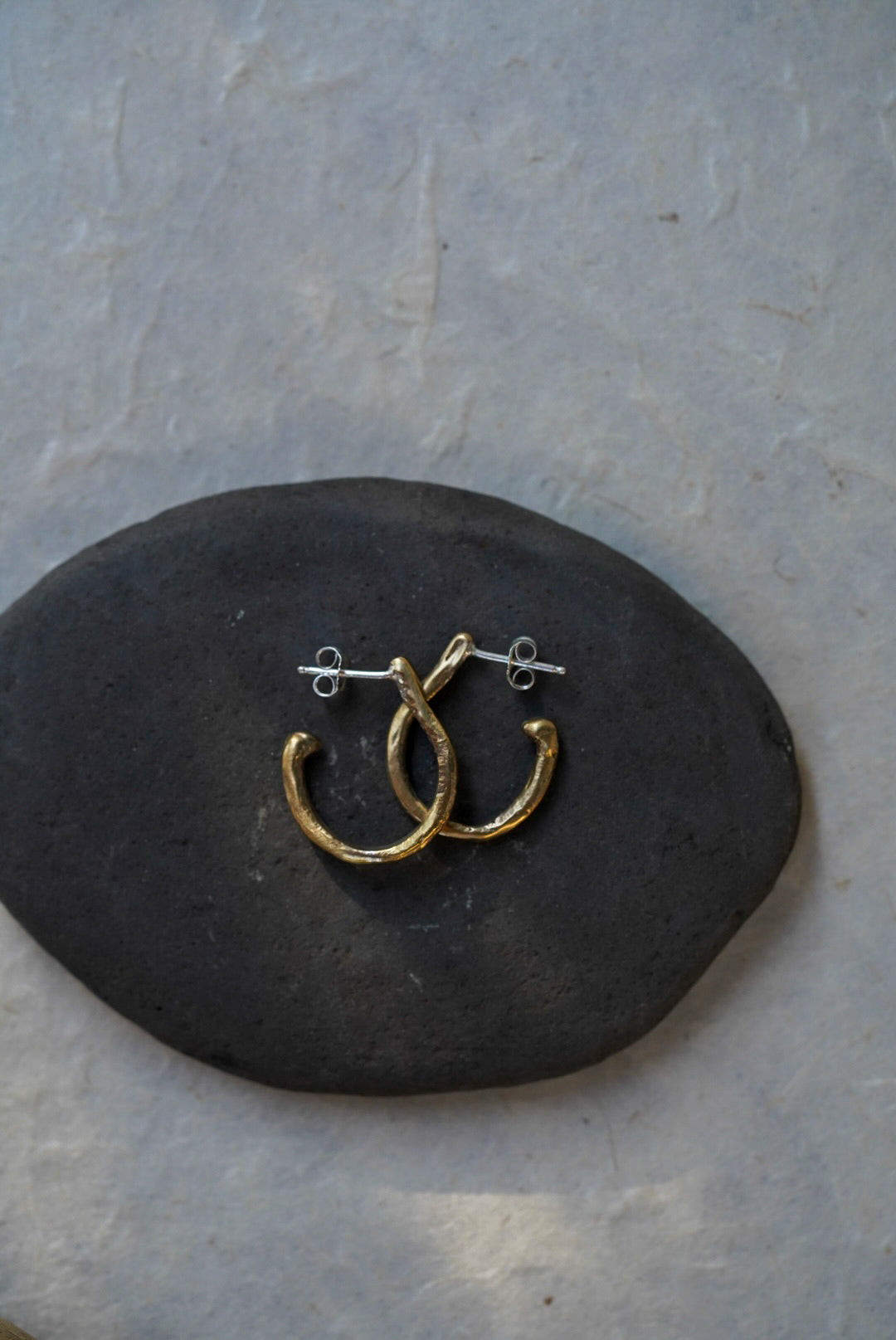 A pair of small bronze hoop earrings with silver posts, displayed on a dark oval stone against a light gray textured background.