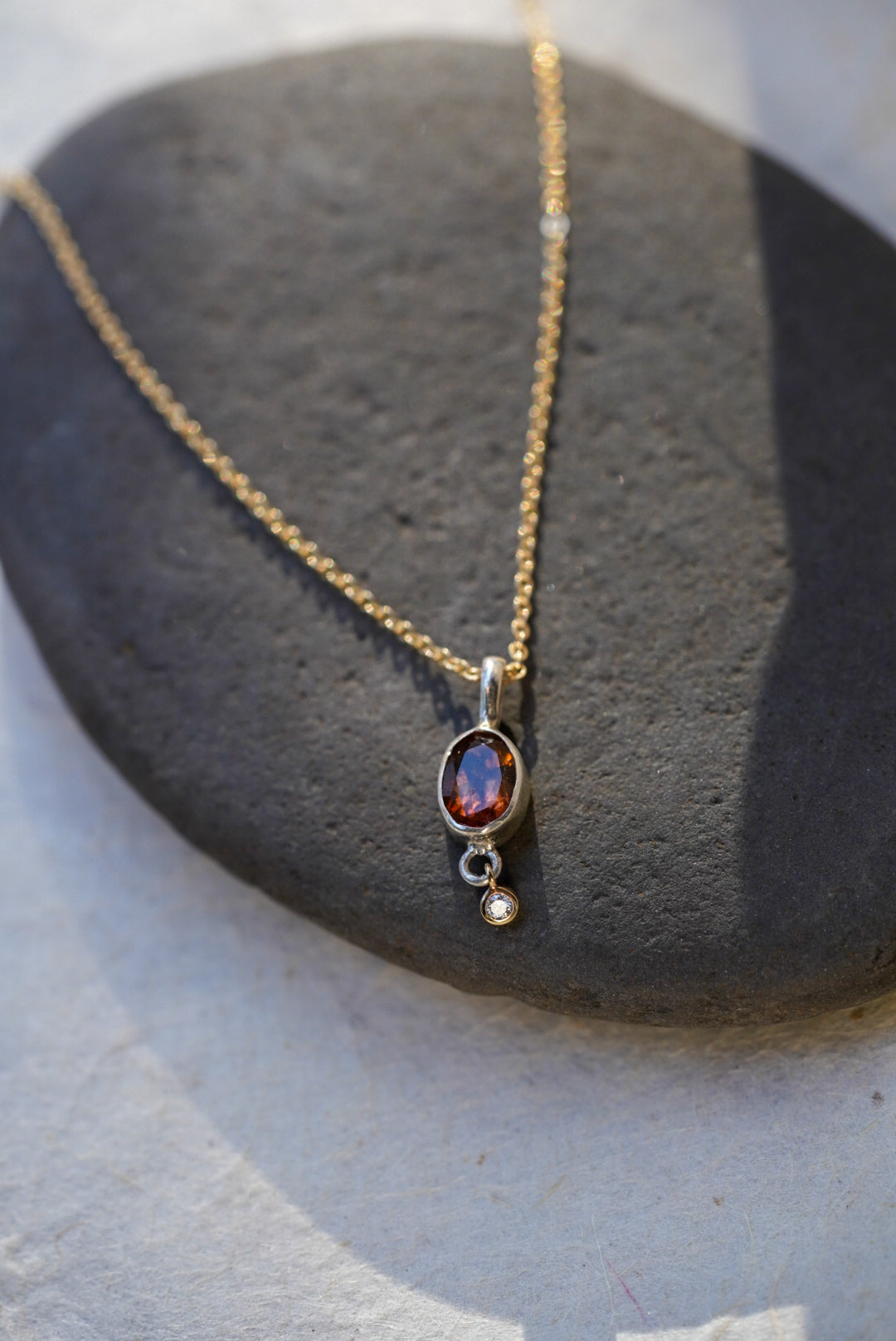 A delicate gold chain necklace with an oval amber-brown gemstone pendant and a small diamond accent stone, displayed on a dark gray stone slab against a light beige background.