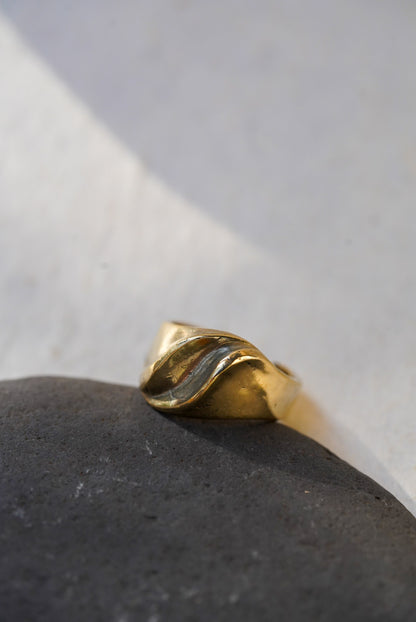 A bronze ring with a twisted, organic wave-like design, resting on a dark textured stone against a light gray background.