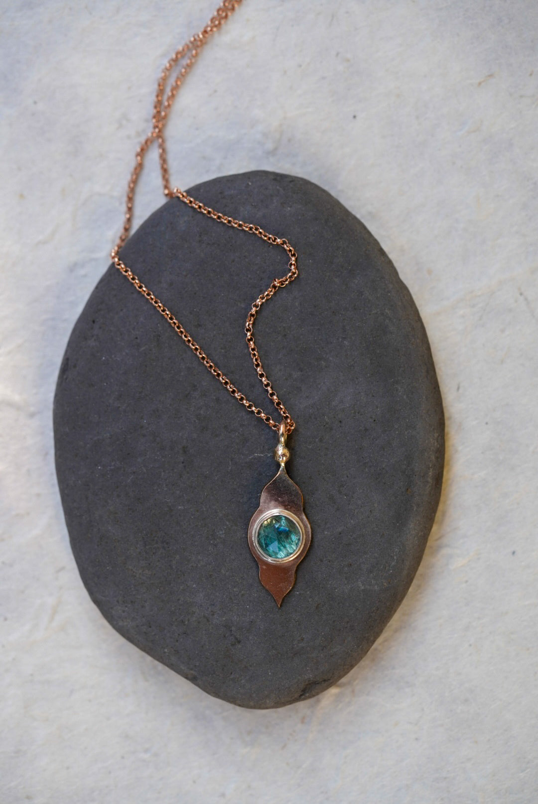 A rose gold necklace with a temple-shaped pendant featuring a round blue-green stone, displayed on a dark slate stone against a light textured background.