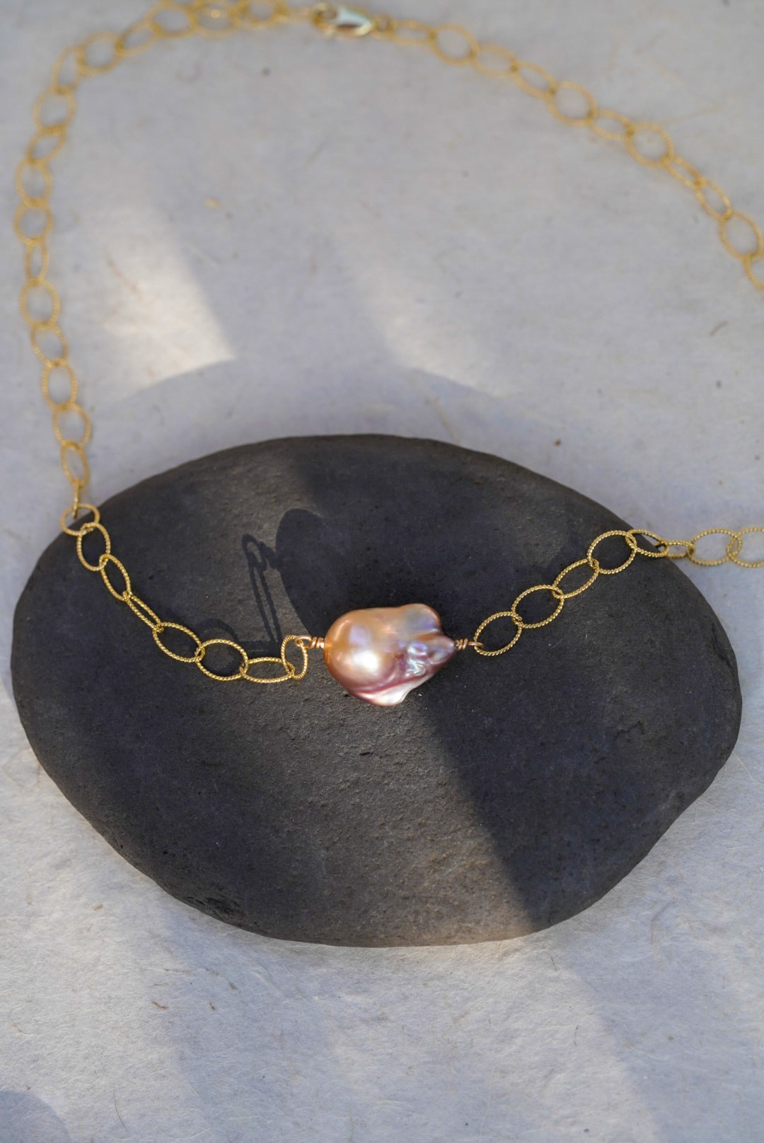 A delicate gold chain necklace featuring a single irregular baroque pearl pendant, displayed on a dark gray stone against a light gray textured background.