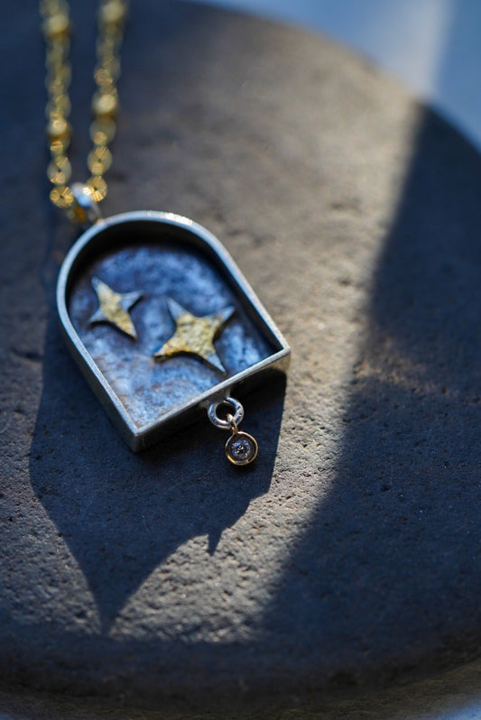 A close-up of a  gold chain necklace with a arched pendant containing two silver stars with a dusting of gold, resting on a dark stone against a textured light background.