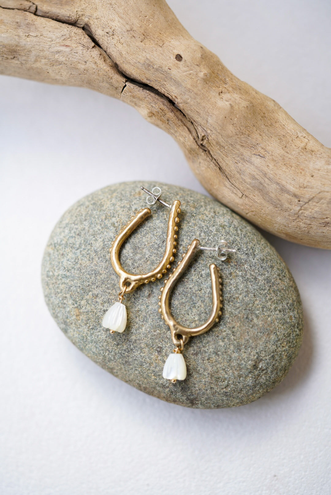 Gold hoop earrings with pearl drops on a stone surface with driftwood.