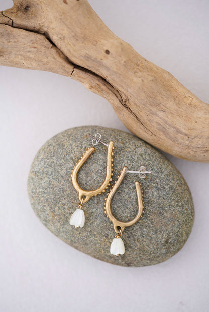 Gold earrings with white pearls on a stone surface with driftwood in the background