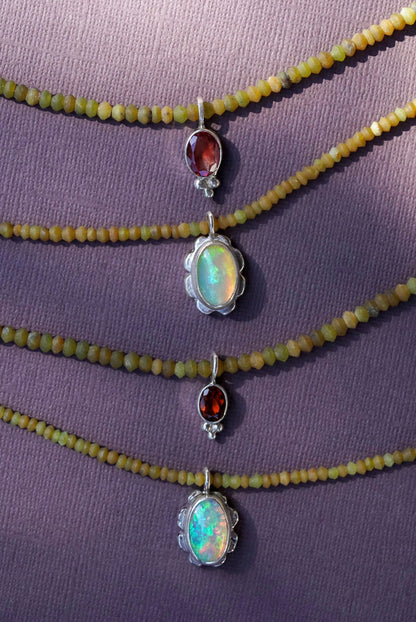 Four beaded strands of olive green beads with silver-toned pendants, including two oval opal-like pendants and two marquise-cut reddish-brown stone pendants, displayed on a textured purple surface.