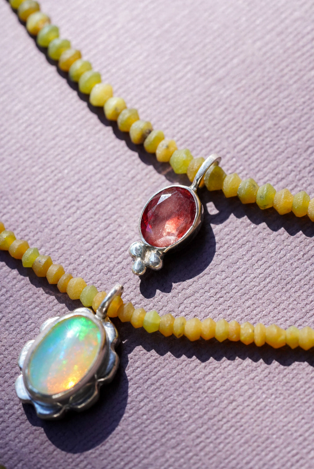 A close-up of a beaded necklace with small yellow beads and three oval pendant settings: one iridescent white opal, one pink gemstone with small clear stones, and one reddish-orange gemstone, all on a textured lavender background.