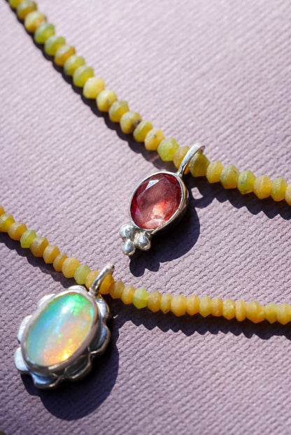 A close-up of a beaded necklace with small yellow beads and three oval pendant settings: one iridescent white opal, one pink gemstone with small clear stones, and one reddish-orange gemstone, all on a textured lavender background.