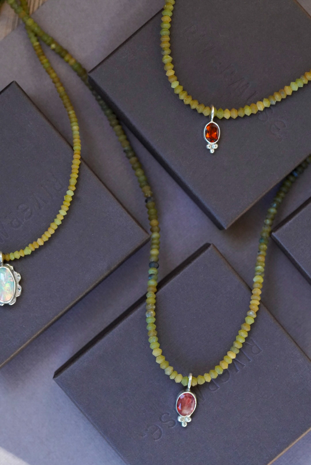 Two beaded necklaces with green-yellow beads and oval red-orange gemstone pendants, displayed on dark brown jewelry boxes.