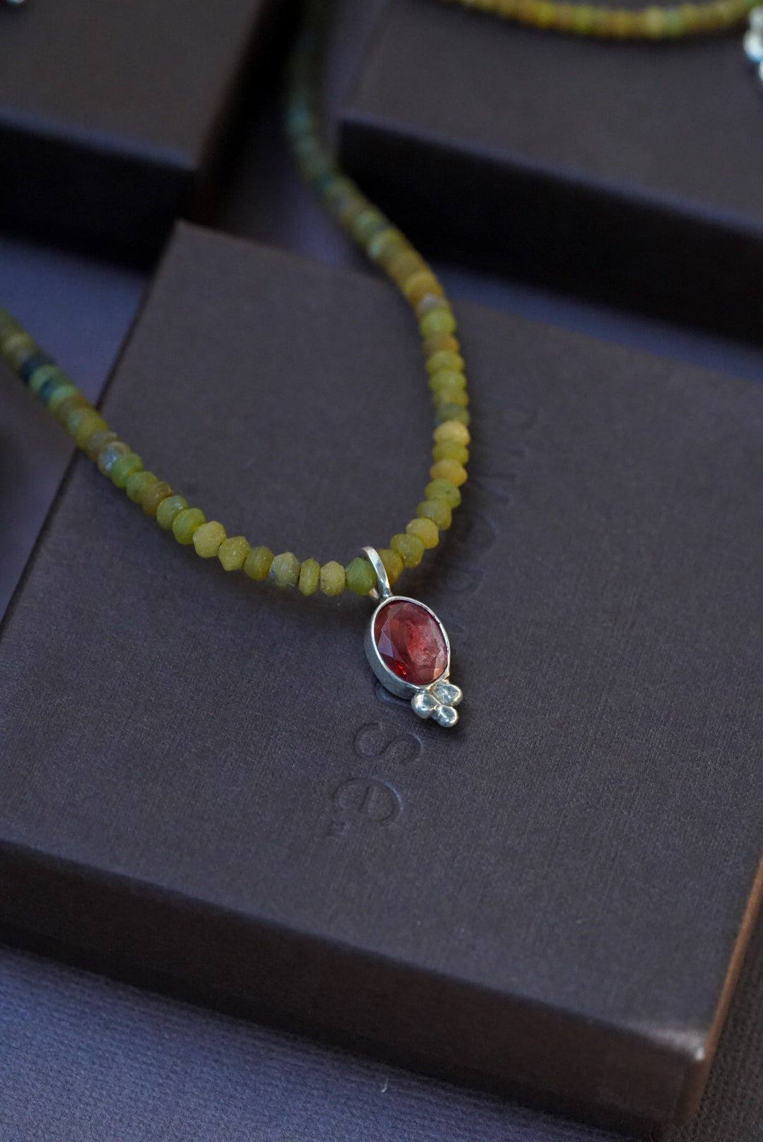 A close-up of a beaded necklace with olive green and yellow beads, featuring a silver pendant with a red oval stone and a small clear gem, displayed on dark brown boxes.