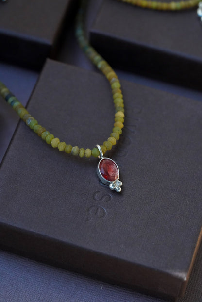 A close-up of a beaded necklace with olive green and yellow beads, featuring a silver pendant with a red oval stone and a small clear gem, displayed on dark brown boxes.