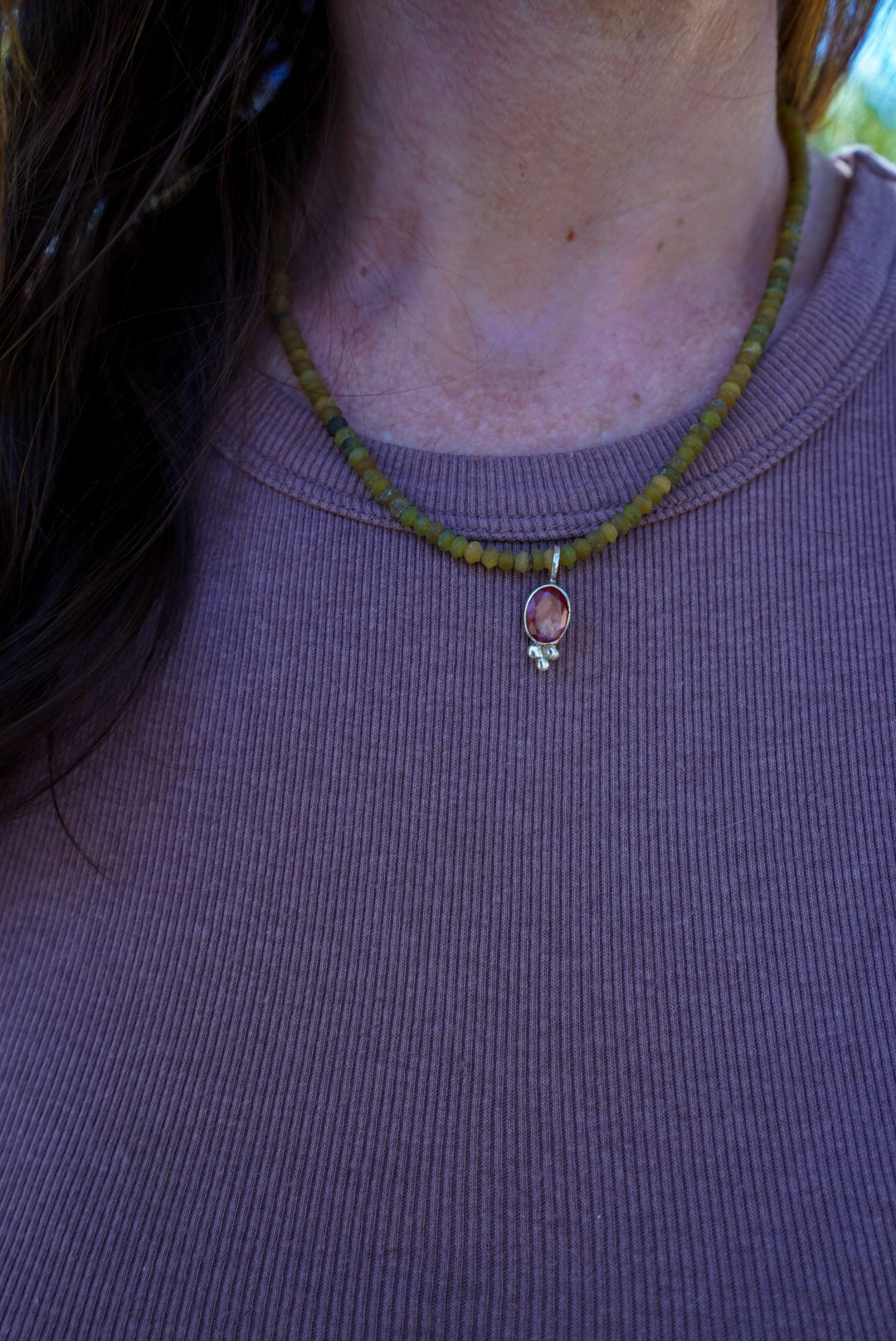 A close-up of a person wearing a beaded necklace with olive green beads and a small pendant, paired with a ribbed brown top.