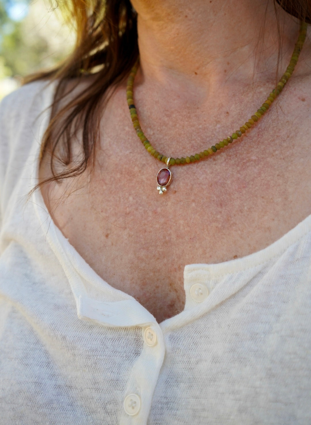 A close-up of a person wearing a beaded necklace with a small oval pendant. The necklace has olive green beads with a small silver set sunstone