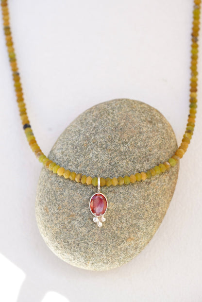 A beaded necklace with olive green and yellow-green beads and a central oval orange sunstone pendant in a silver setting, displayed on a smooth gray stone against a white background.
