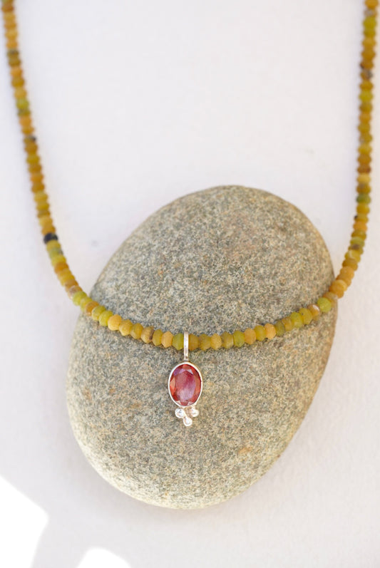 A beaded necklace with olive green and yellow-green beads and a central oval orange sunstone pendant in a silver setting, displayed on a smooth gray stone against a white background.