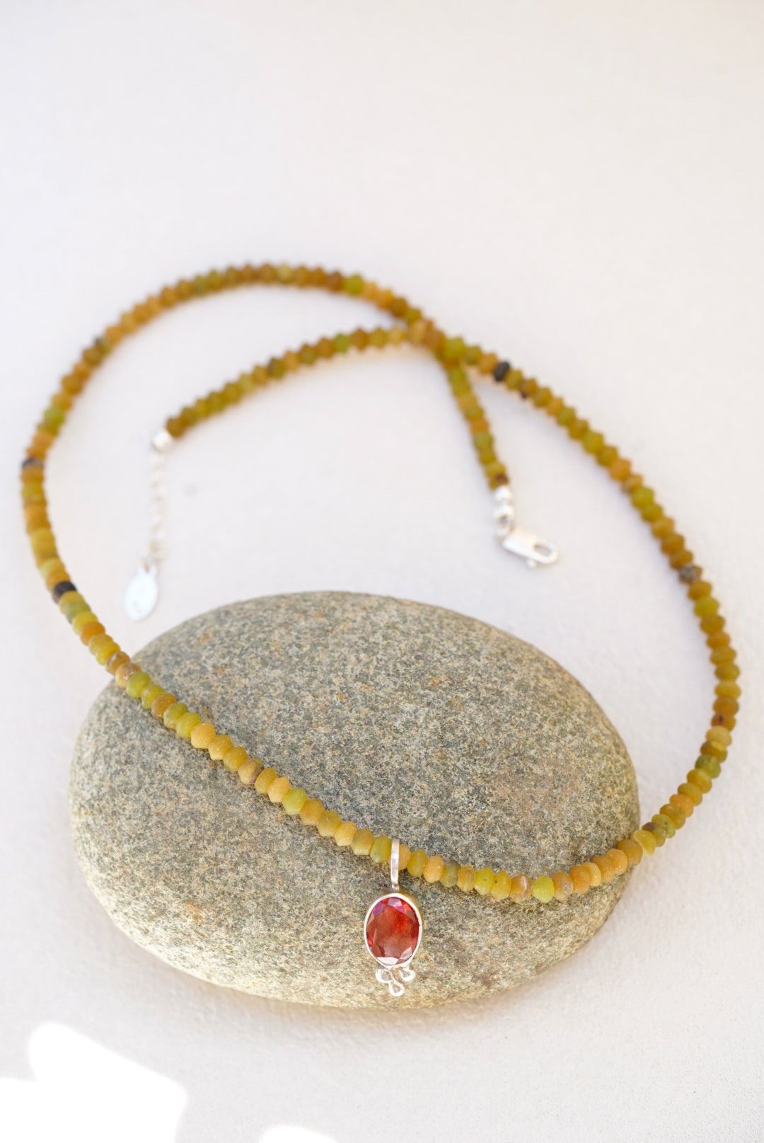 A beaded necklace with olive green and yellow-green round beads, featuring a small oval red-orange stone pendant in a silver bezel, displayed on a smooth gray stone against a light background.