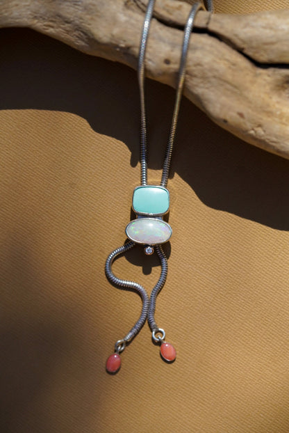 A silver snake-chain bolo necklace with a pendant featuring a light blue stone, a white opal, and two dangling coral-pink beads, displayed on a tan textured surface with a piece of driftwood.