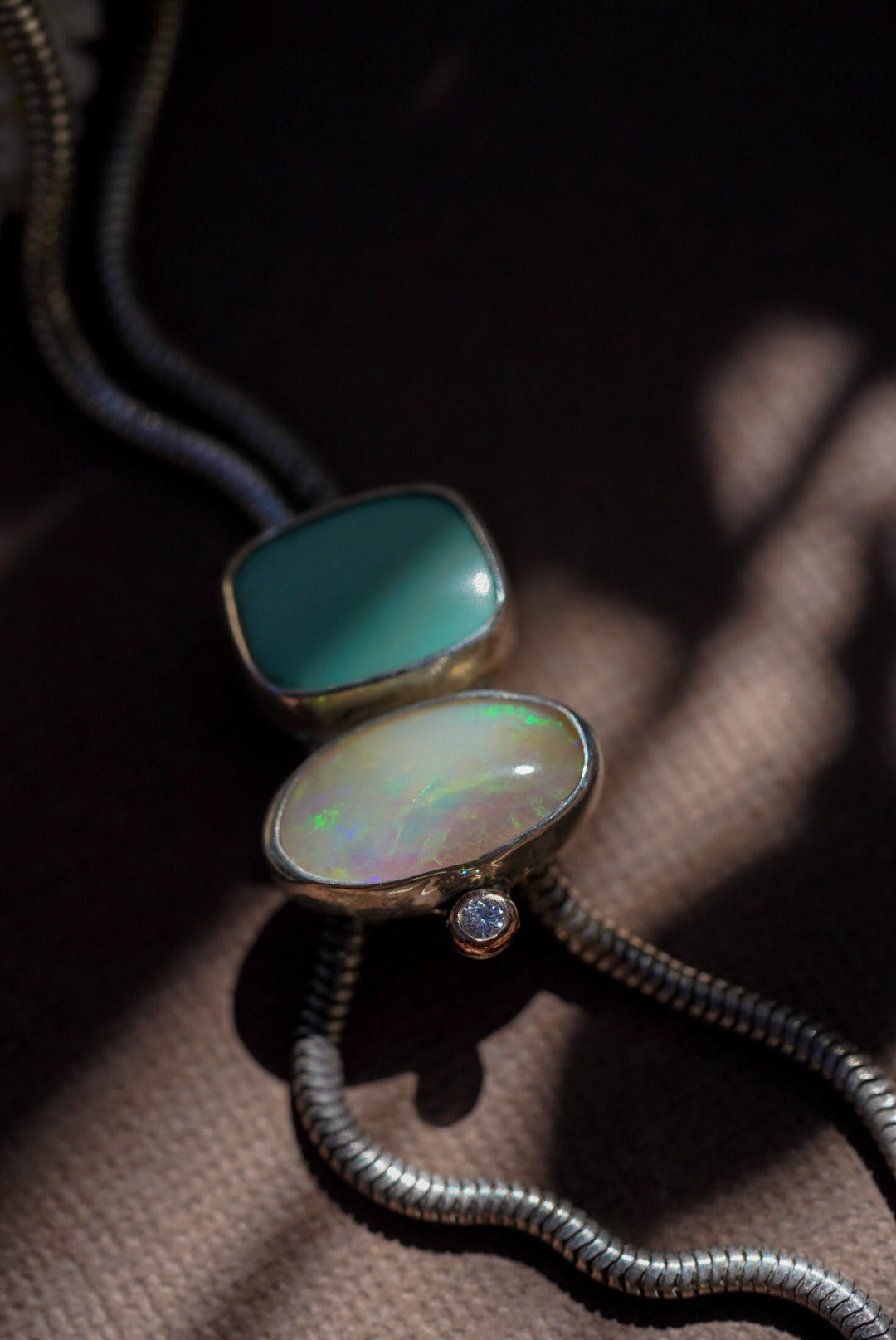 A close-up of a silver snake chain bolo necklace featuring two stones, one with a smooth teal-green finish and the other with a multicolored iridescent opal, resting on a textured brown surface.