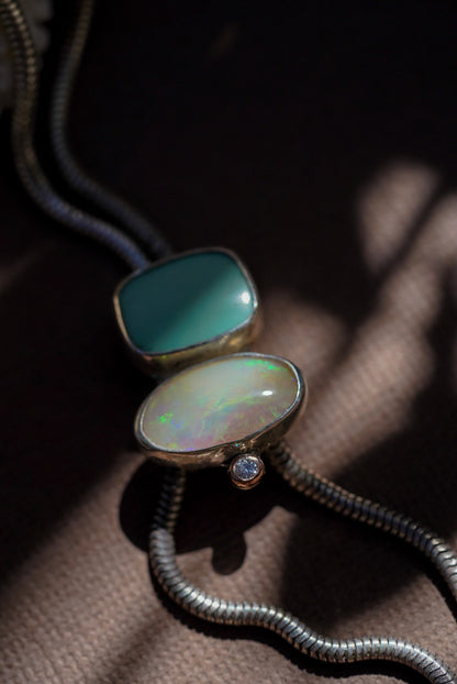 A close-up of a silver snake chain bolo necklace featuring two stones, one with a smooth teal-green finish and the other with a multicolored iridescent opal, resting on a textured brown surface.