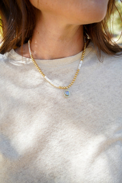 A close-up lifestyle shot of a person wearing a delicate gold beaded necklace with a small oval opal stone pendant, paired with a white pearl and gold beaded necklace.
