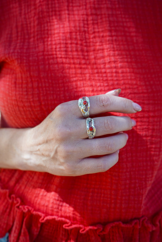 A close-up of a person's hand wearing two silver rings with red coral accents, set against a vibrant red textured fabric background.