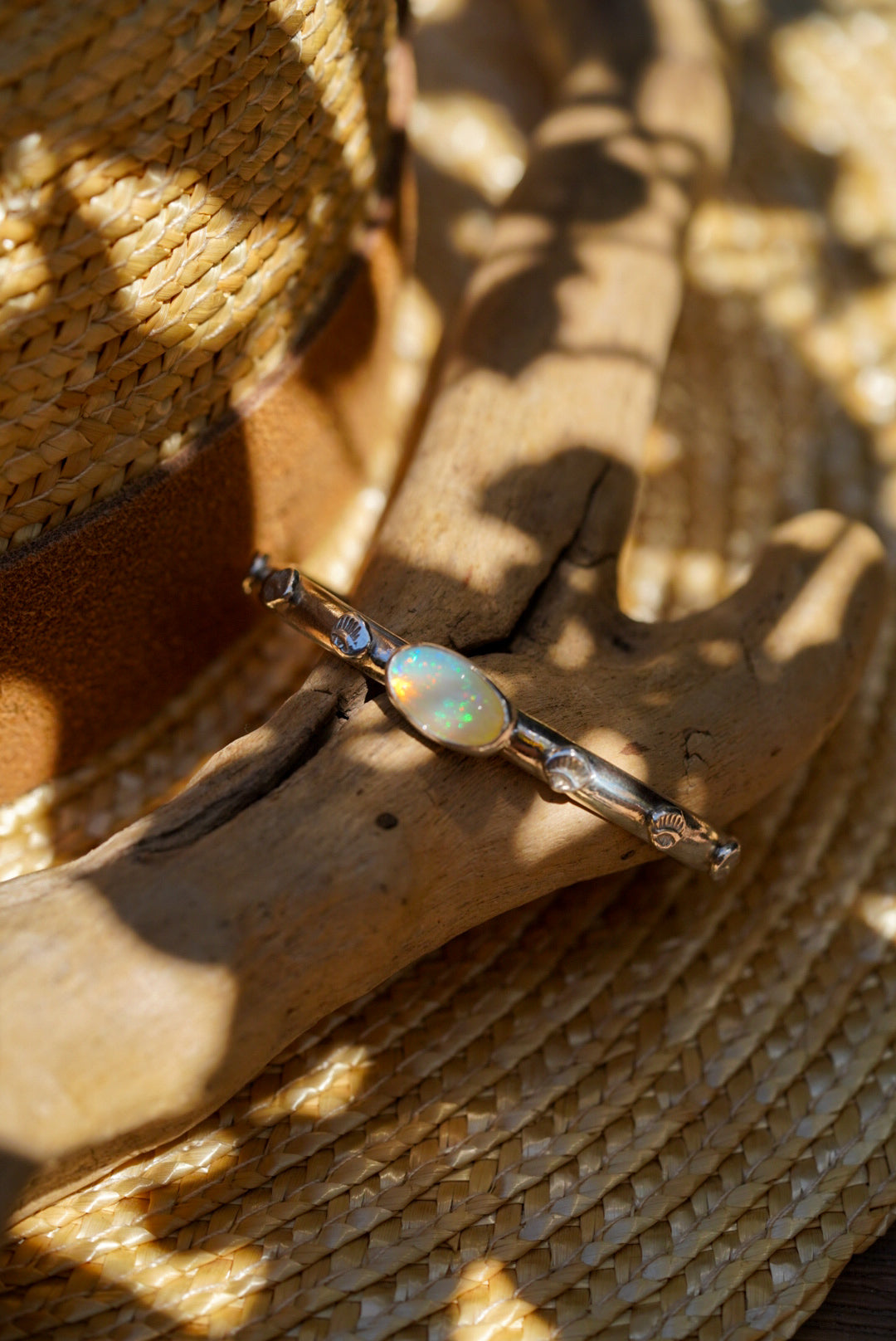 A silver bracelet with a central iridescent opal stone, displayed on a piece of driftwood resting on a woven straw hat.